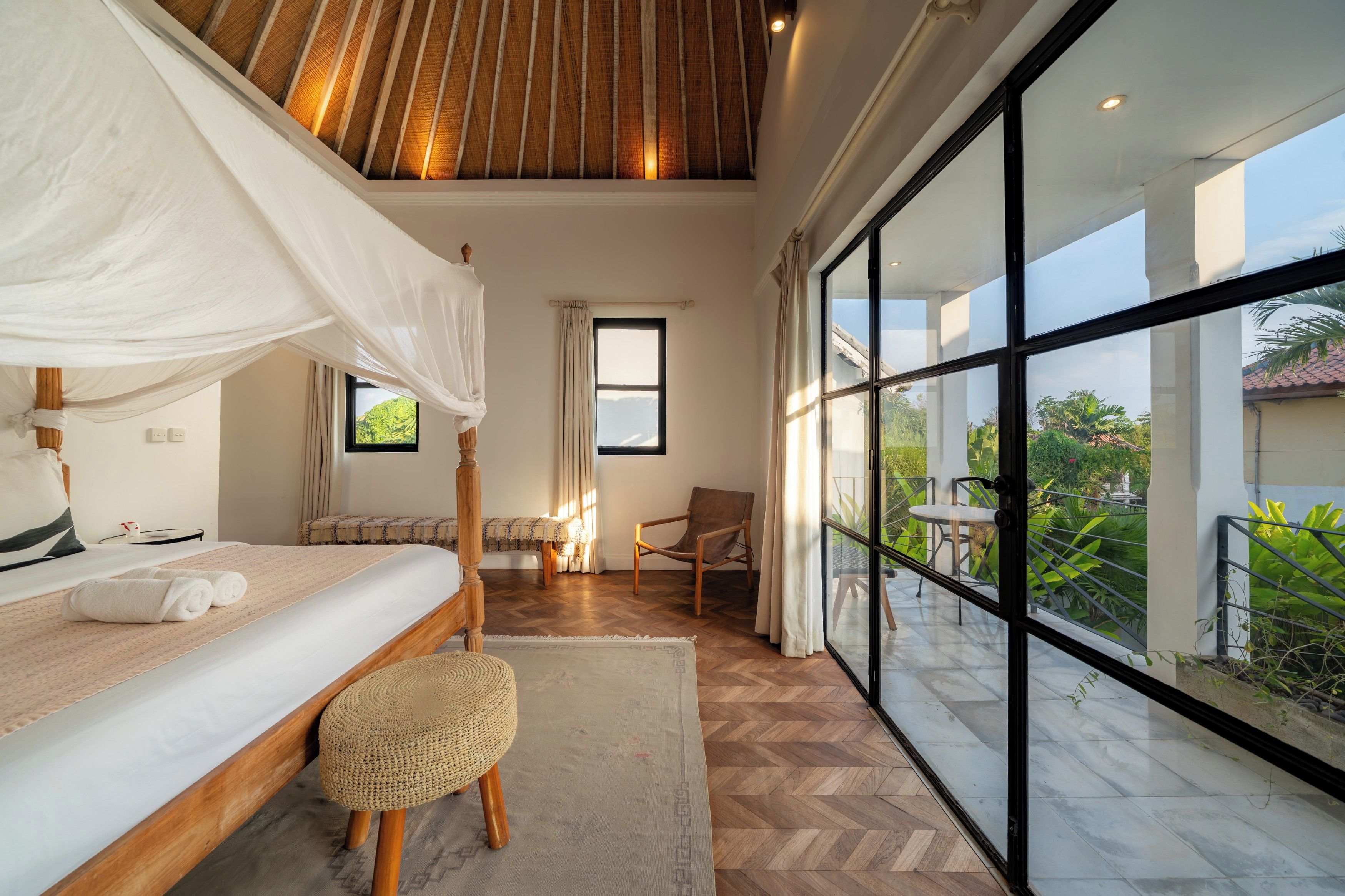Ein Schlafzimmer mit Baldachinbett, einer Sitzecke mit Sessel und Tisch sowie einem Balkon mit Blick auf Grünflächen.