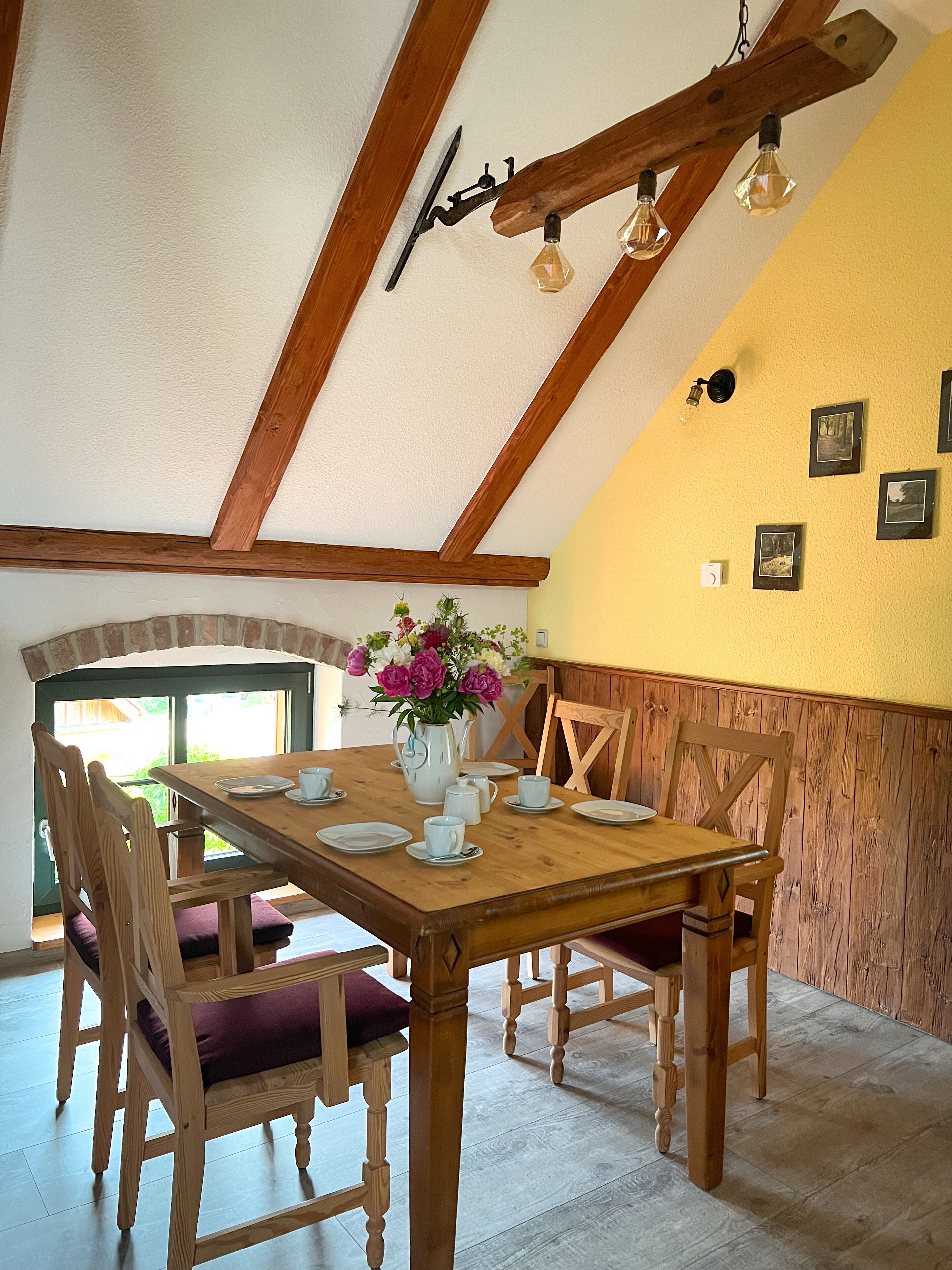 Un comedor con una mesa de madera dispuesta para cuatro personas, rodeada de sillas de madera con cojines morados. Un jarrón con flores está sobre la mesa, y hay cuadros enmarcados en la pared.