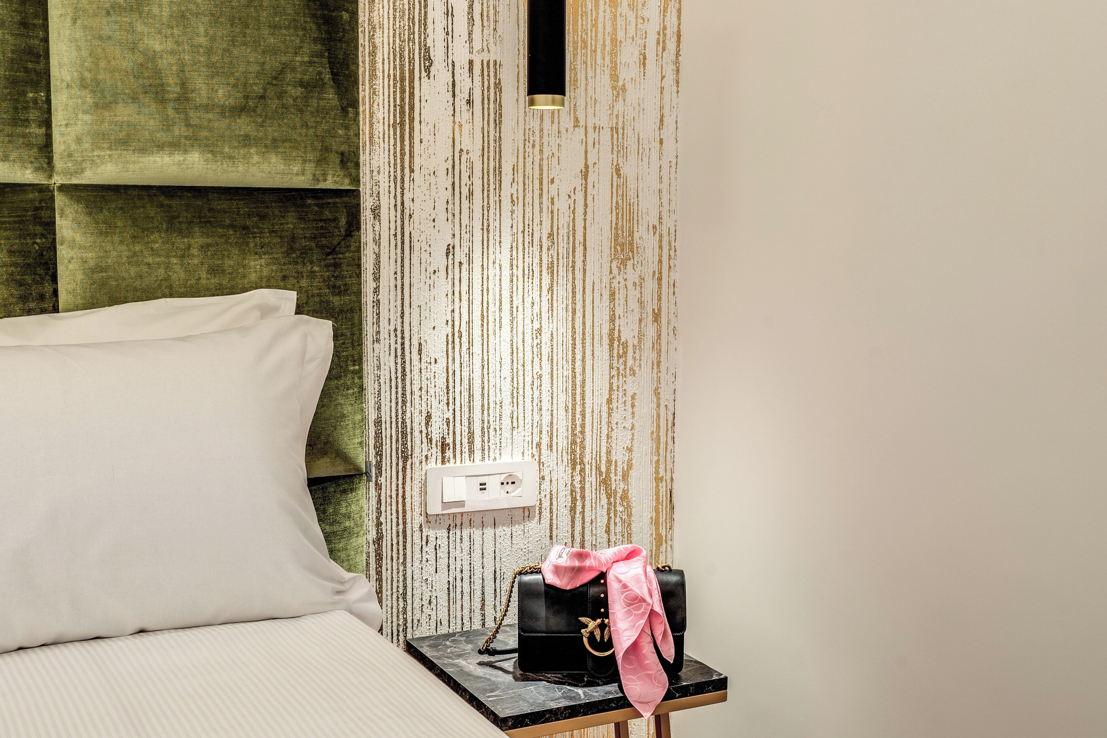 A neatly made bed with a white pillow, a black handbag with a pink scarf on a small table, and a power outlet on the wall.