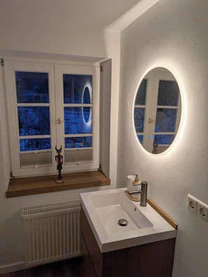 Une salle de bain avec un lavabo blanc, un miroir et une fenĂȘtre donnant sur des arbres.