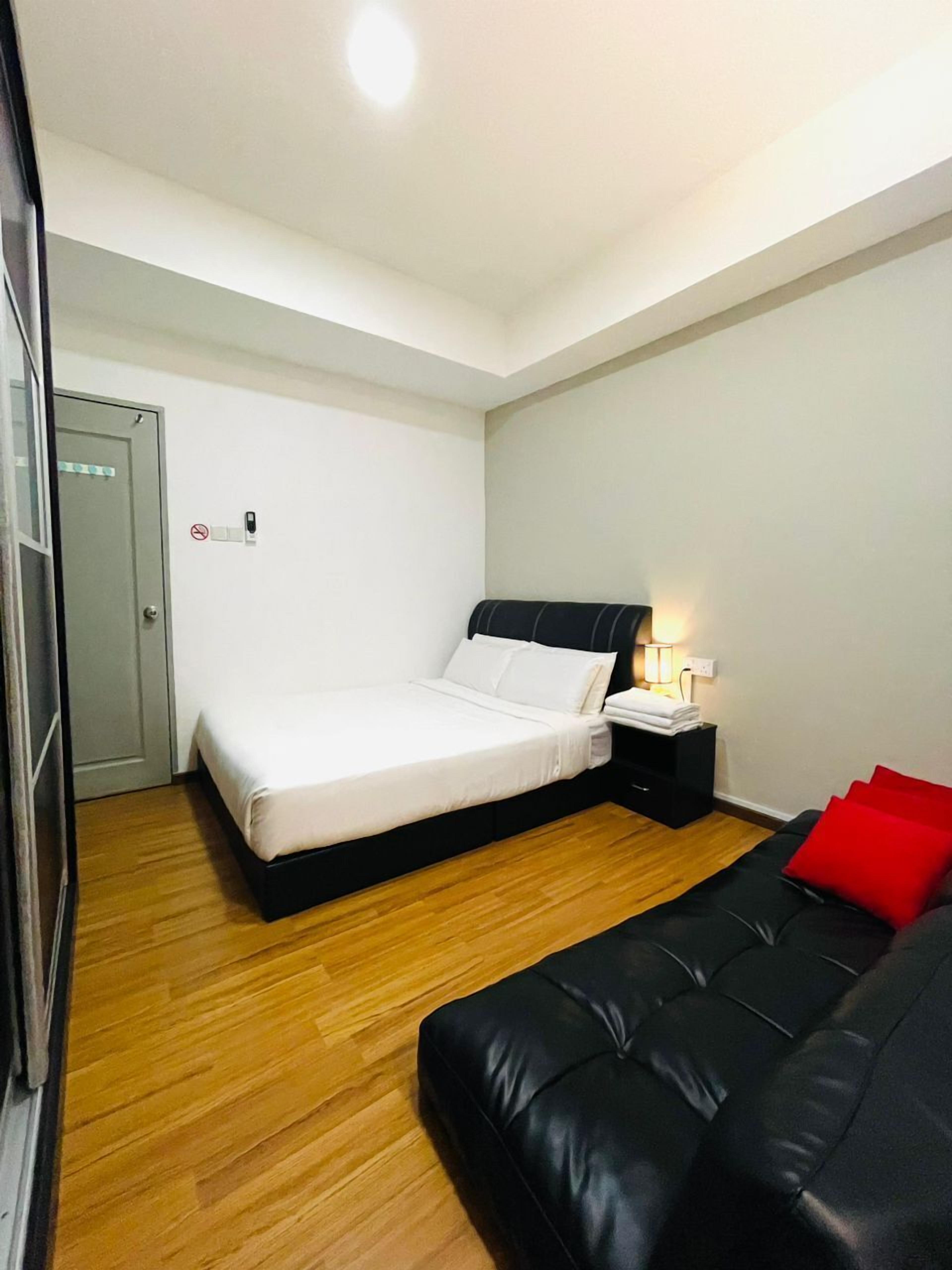 Un dormitorio con cama, mesita de noche, almohada roja, sofá negro y suelo de madera.