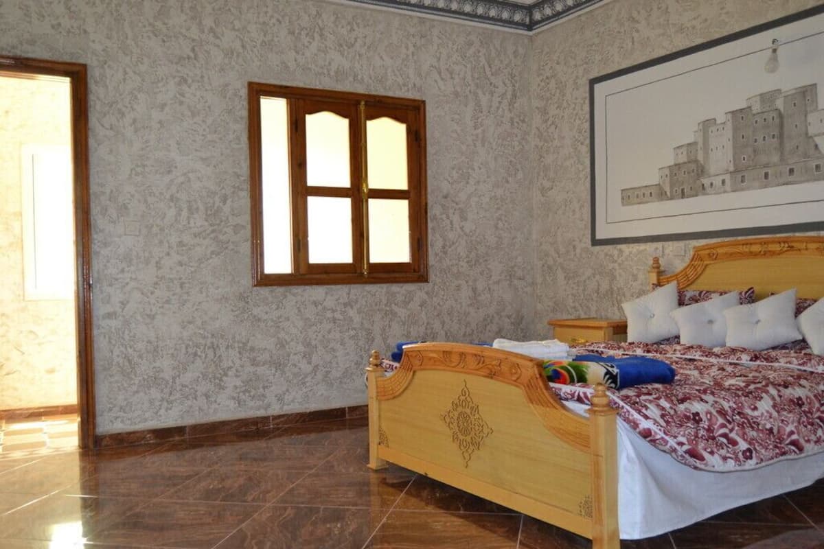 A bedroom with a wooden bed, a nightstand, a framed picture on the wall, and a window with wooden shutters.
