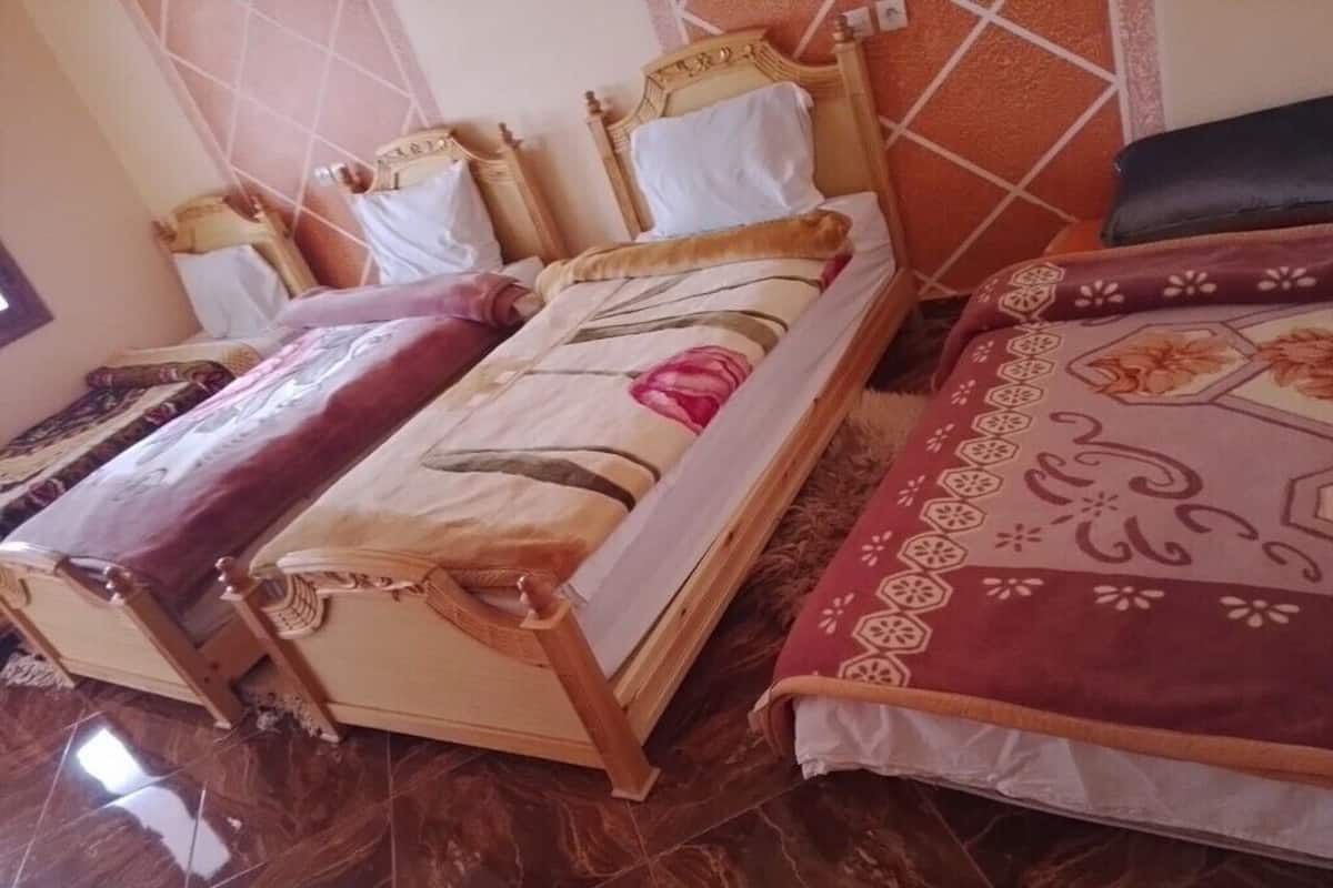 A room with two beds, each with a wooden frame and headboard. The beds have white and patterned bedding. The room has a tiled floor.