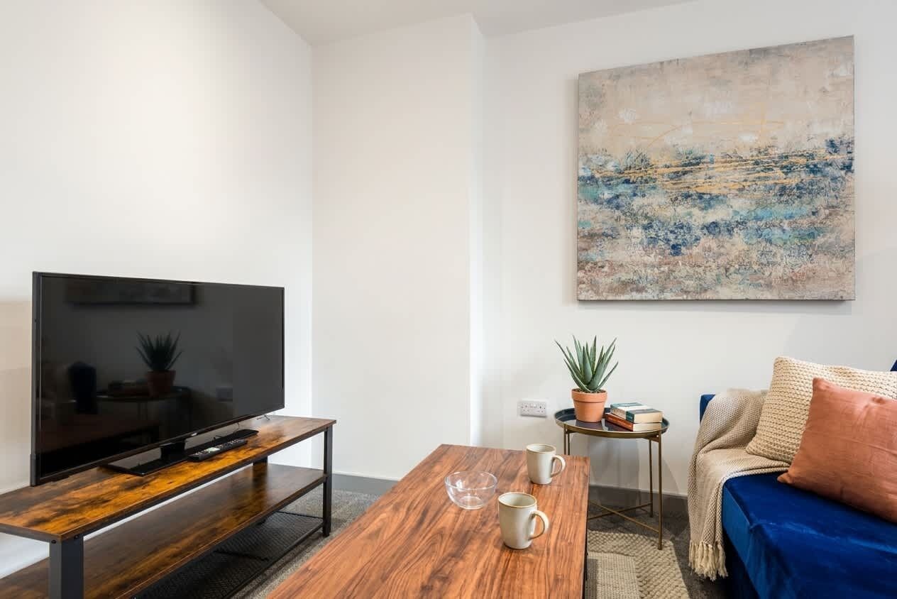 Una sala de estar moderna con un televisor de pantalla plana sobre un soporte de madera, una mesa de centro con dos tazas, un sofá con un cojín y un cuadro en la pared.