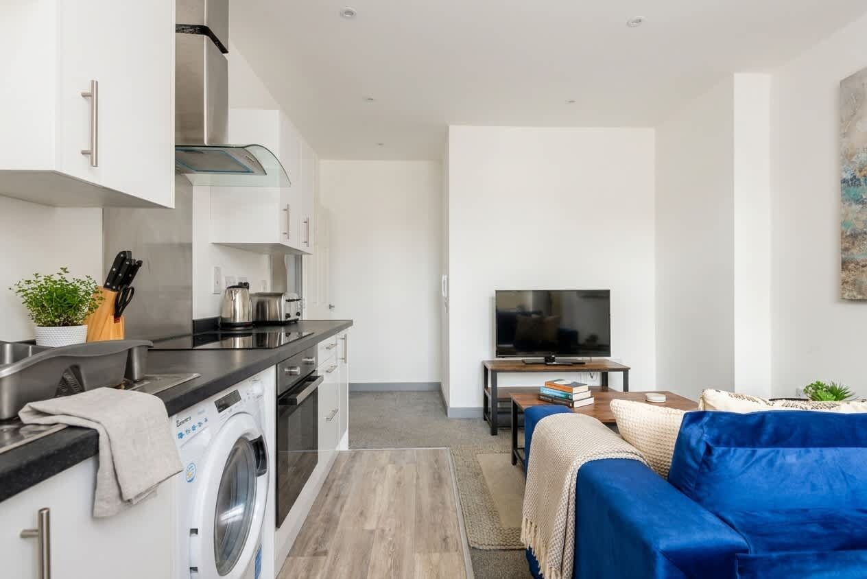 A modern kitchen with a washing machine, a dining area with a blue sofa, and a TV.