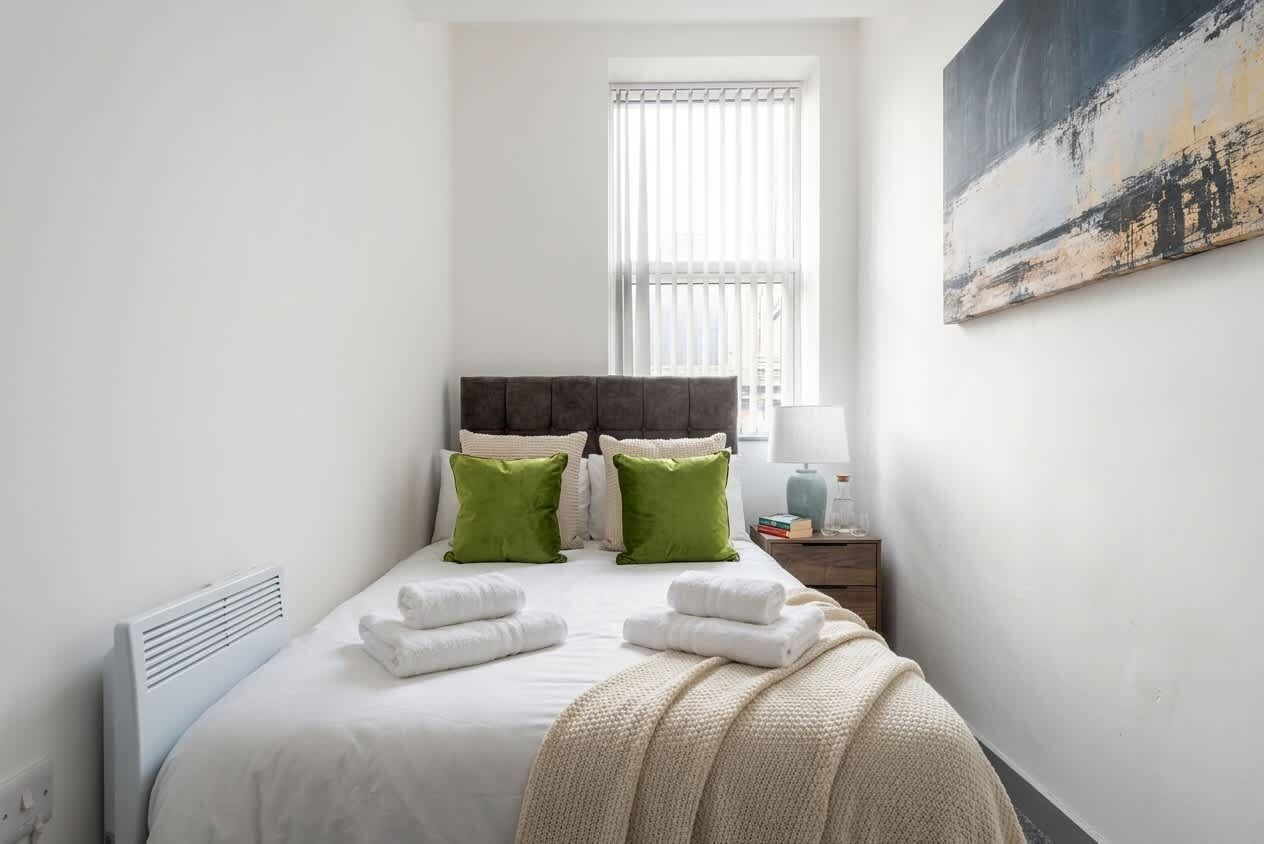 A bedroom with a bed, pillows, a radiator, a window, and a painting on the wall.