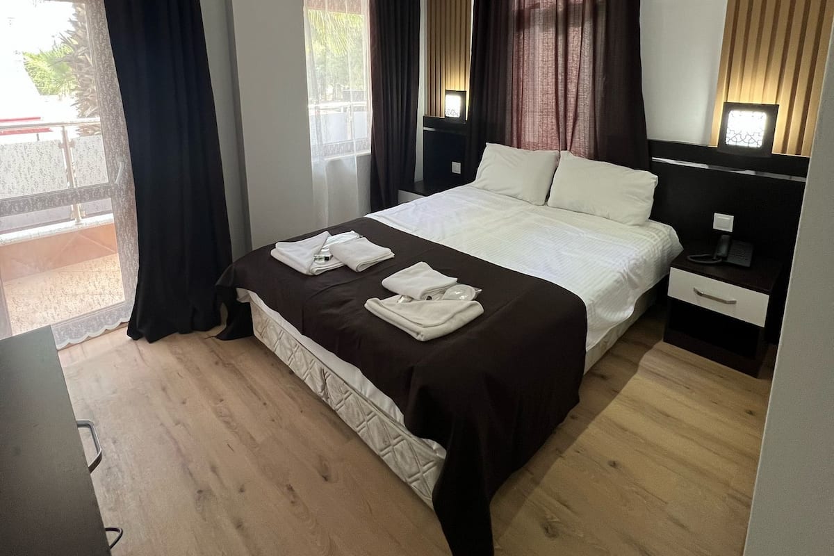 Une chambre d’hôtel moderne avec un grand lit, une table de chevet, un bureau et une chaise. On y trouve une fenêtre avec des rideaux, un luminaire au plafond et un climatiseur fixé au mur.