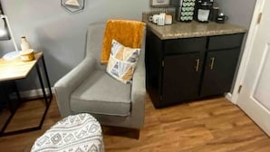 A room with a grey armchair, a patterned ottoman, a hexagonal wall decoration, a dark cabinet with gold handles, and a small table with a lamp.