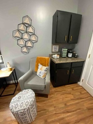 Un petit salon moderne avec un canapé gris, un pouf à motifs, une table basse, un luminaire hexagonal fixé au mur et des meubles sombres dotés de poignées dorées.