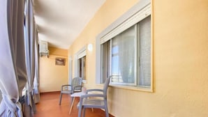 Un pasillo con dos sillas, una mesa, una ventana con mosquitero y una puerta con lámpara.