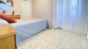Un dormitorio con cama, mesita de noche, armario y una pequeña mesa con lámpara.