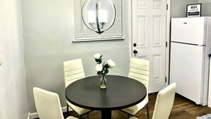 Un comedor con una mesa redonda, sillas blancas, un jarrón con flores, una lámpara colgante, una puerta blanca y un refrigerador blanco.