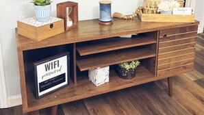 Un mueble de madera para entretenimiento con un televisor de pantalla plana, una planta pequeña, objetos decorativos y un cartel con la contraseña de WiFi.