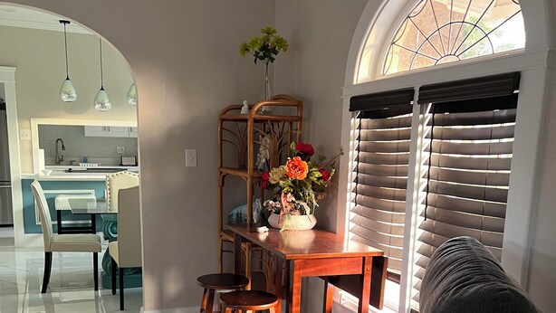 Una sala de estar con un sofá de color gris oscuro, una mesa de comedor de madera con sillas, un jarrón con flores y una ventana con persianas.