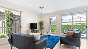 A modern living room with a flat-screen TV, two sofas, a coffee table, and large windows offering a view of the outdoors. - Your family & staff will love it here! Bed & bath linen hire included. Unlimited WIFI. Free, easy parking. Free child friendly amenities. Keyless access. Easy self check in. No stairs! (Richmond)