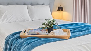 A neatly made bed with a blue blanket, a wooden tray with a book and a plant, and a painting of a sunset on the wall. - Your family & staff will love it here! Bed & bath linen hire included. Unlimited WIFI. Free, easy parking. Free child friendly amenities. Keyless access. Easy self check in. No stairs! (Richmond)