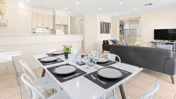 A modern kitchen with white cabinets, a dining area with a table set for four, and a grey sofa in the living area. - Your family & staff will love it here! Bed & bath linen hire included. Unlimited WIFI. Free, easy parking. Free child friendly amenities. Keyless access. Easy self check in. No stairs! (Richmond)