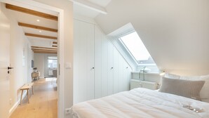 Ein Schlafzimmer mit Dachfenster, Holzbalken, einem Bett sowie Blick auf eine Küche und ein Badezimmer.