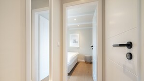 Ein Flur, der in ein Schlafzimmer mit einem Bett, einem kleinen Hocker und einem gerahmten Bild führt.