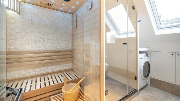 Eine Sauna mit Holzbänken und einem Holzfass, ein abgeschlossener Duschbereich und eine Waschmaschine in einem kleinen Raum mit Dachfenster.