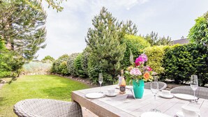 Ein Essplatz im Freien mit weißen Tellern, Gläsern und einer Flasche Alkohol, umgeben von Korbstühlen und einem Blumenstrauß.
