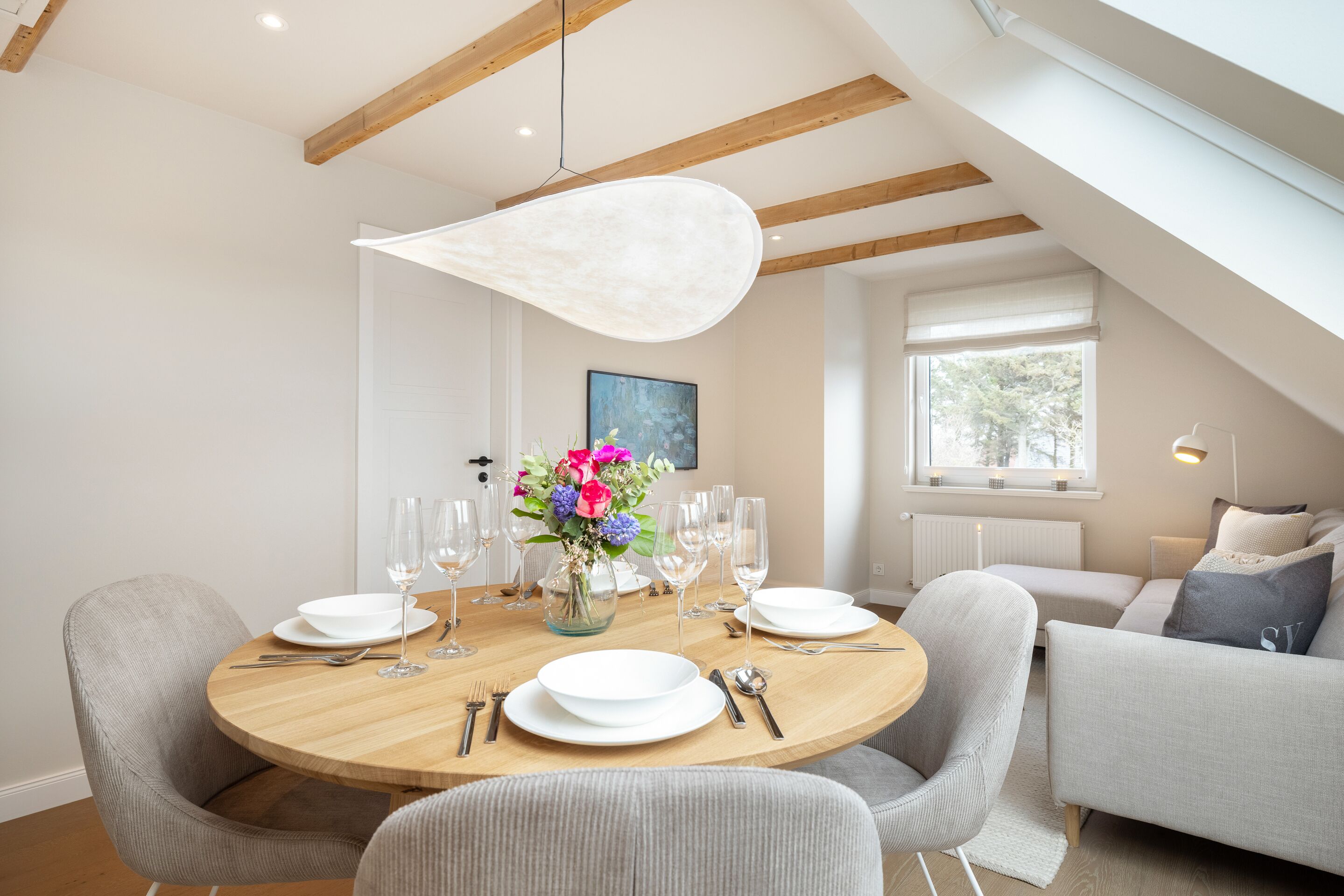 Un coin repas avec une table ronde dressée pour six personnes, entourée de chaises à revêtement gris. Un grand lustre est suspendu au-dessus de la table, et un vase rempli de fleurs est placé au centre.