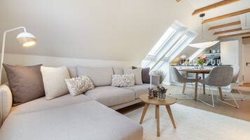 Un appartement moderne de style « loft », doté d’un grand canapé en forme de L, d’un coin repas avec une table et des chaises, et d’une cuisine située à l’arrière-plan.