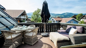 A terrace with wicker furniture, a table with cups and a teapot, and a view of houses and mountains.