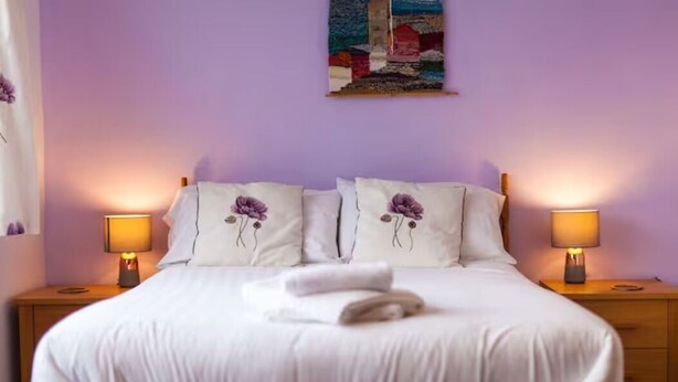 A neatly made bed with white linens and two pillows, each with a purple floral design. There are two bedside lamps on wooden nightstands, and a colorful painting hangs on the wall above the bed. - Oak House (Machynlleth)