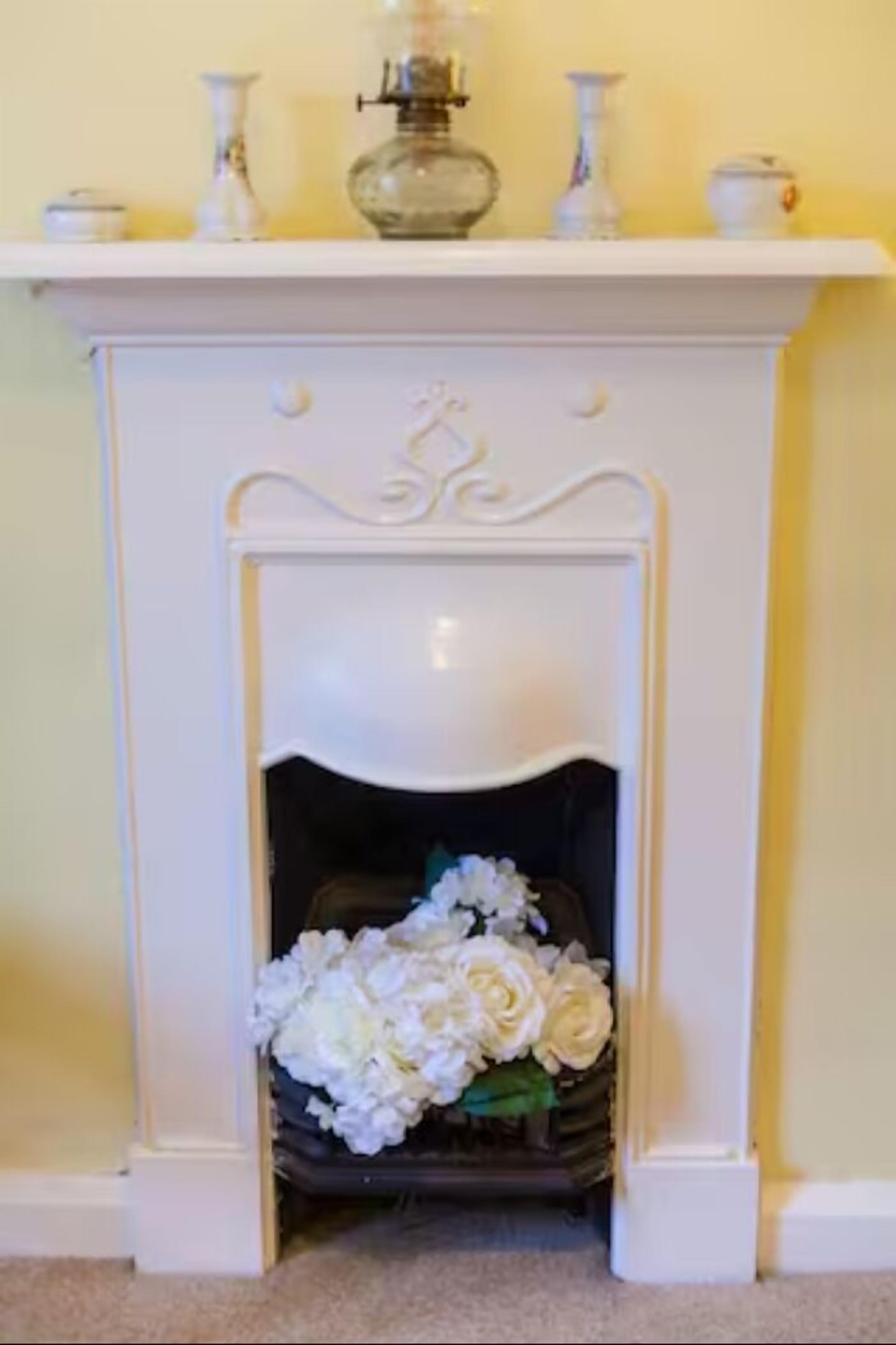 Una chimenea blanca con un estante decorativo en la parte superior y una cesta de flores blancas abajo.