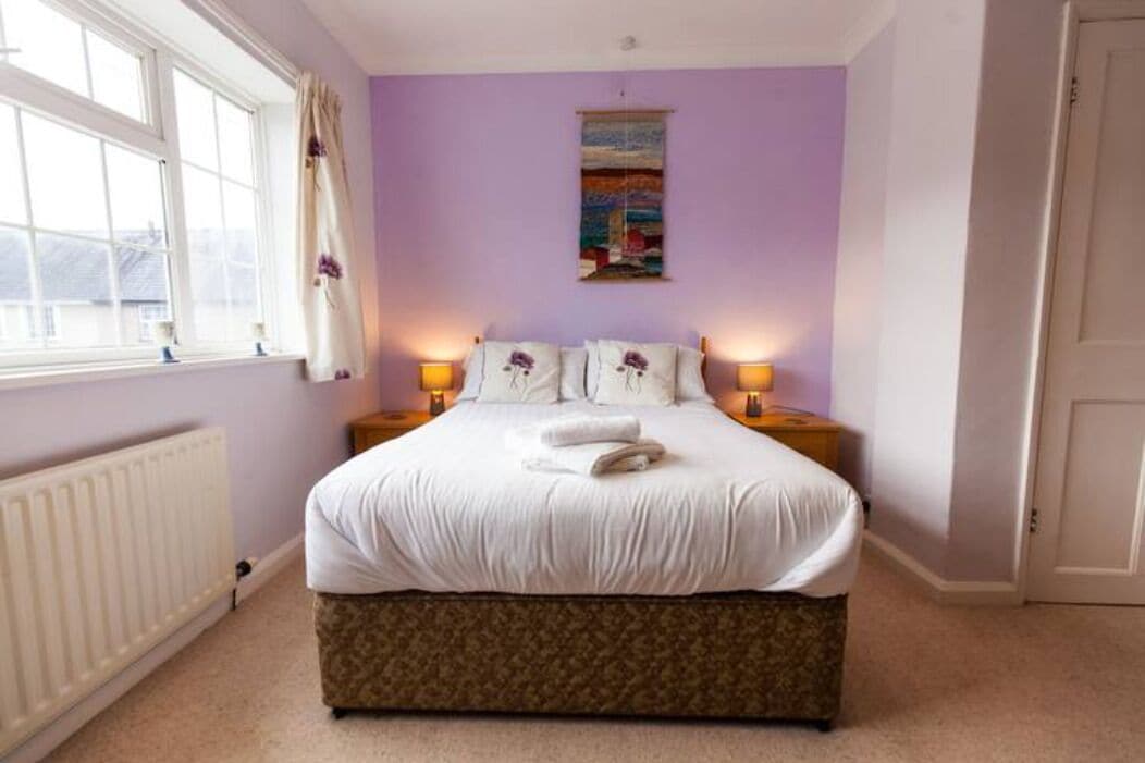 A bedroom with a large bed, two bedside tables with lamps, a window with curtains, a radiator, and a painting on the wall.