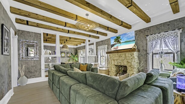 A spacious living room with a green sectional sofa, wooden beams on the ceiling, a stone fireplace, and a large window with curtains. - Casa Alaïa | West Brickell | Sleeps 20 | Pool&Spa (Miami)