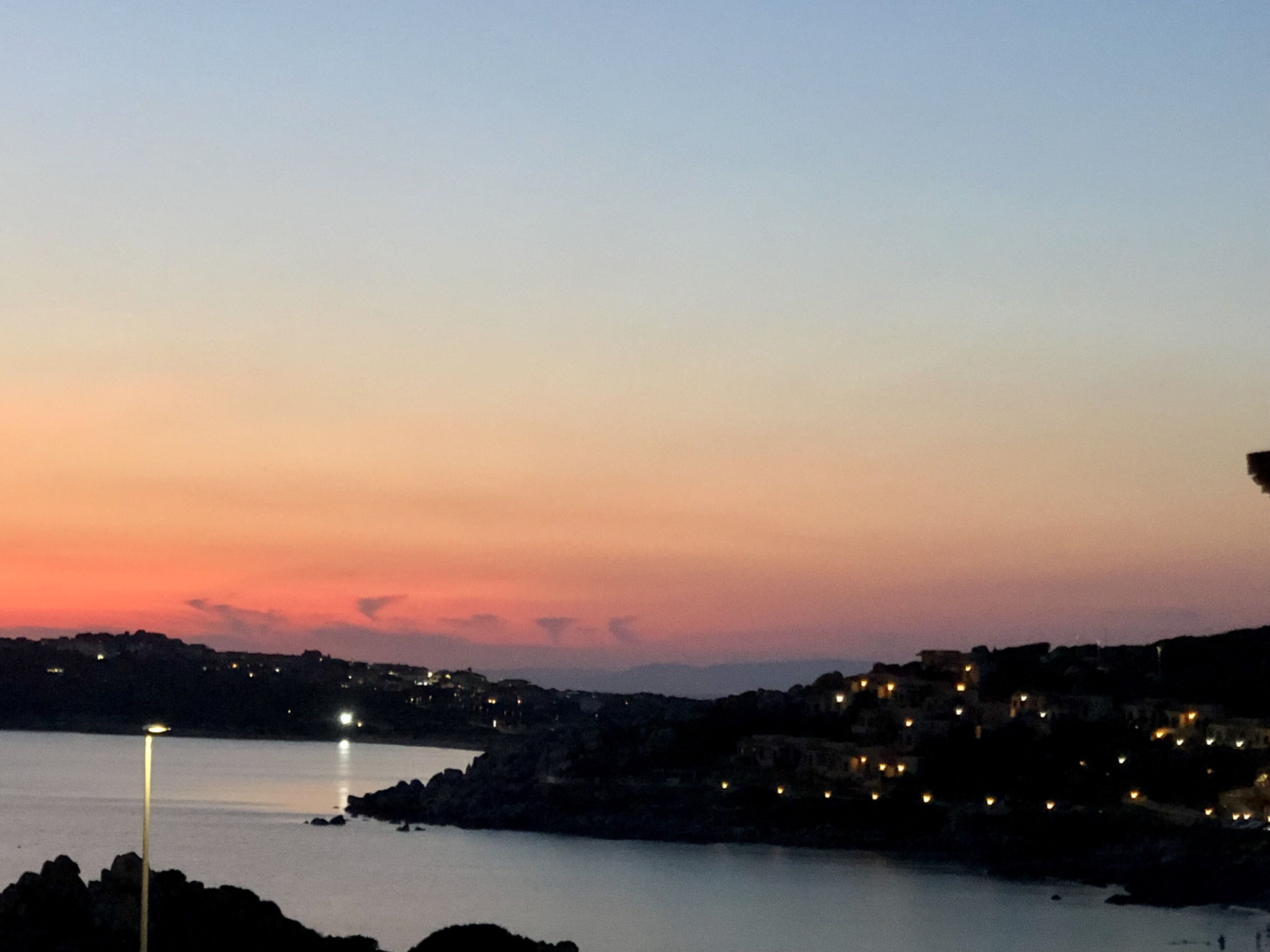 A coastal town at dusk with a calm sea, a prominent street lamp, and buildings with lights on.