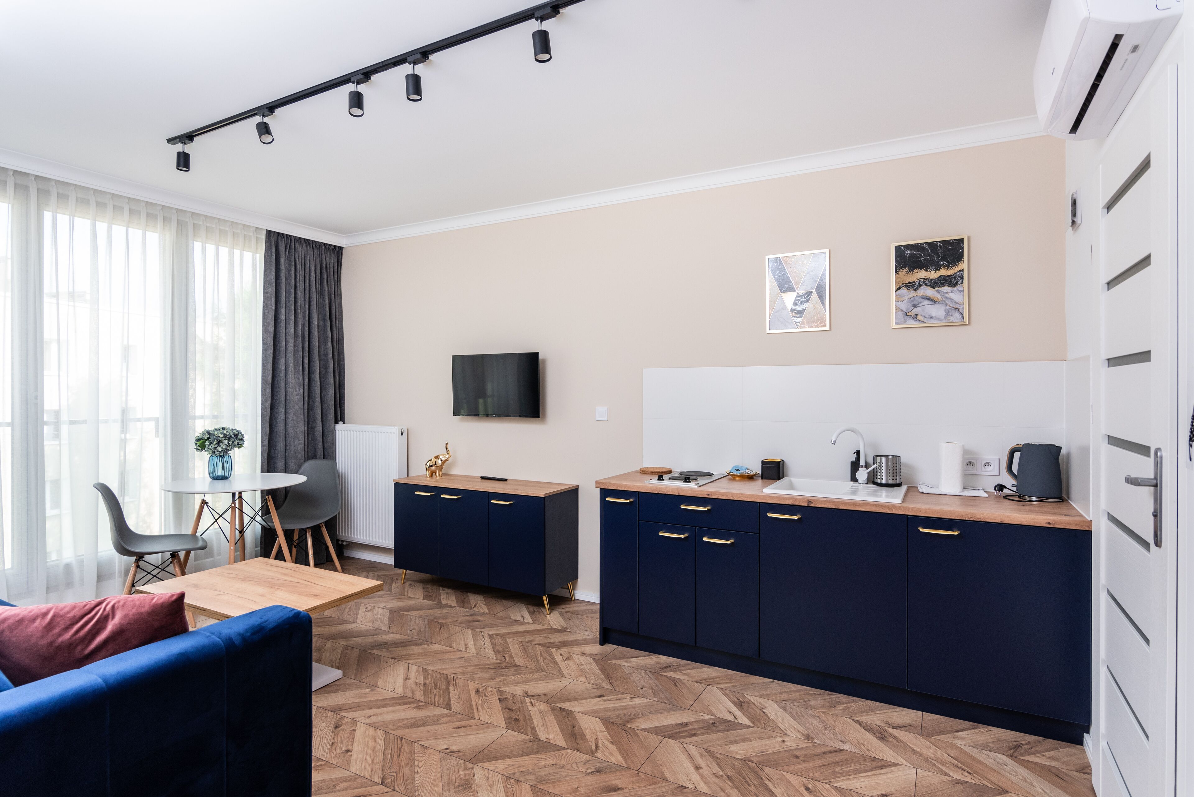 Un salon moderne avec un canapé, une table basse et un coin repas avec des chaises. On y trouve une cuisine avec des meubles bleu foncé et un plan de travail en bois. Un téléviseur à écran plat est fixé au mur, et deux tableaux encadrés sont accrochés au-dessus du plan de travail.