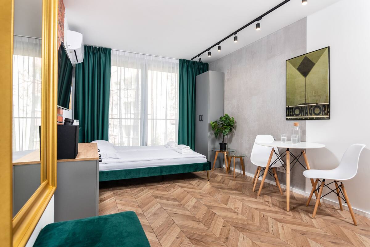 A modern hotel room with a bed, a desk with chairs, a potted plant, and a wall-mounted artwork.