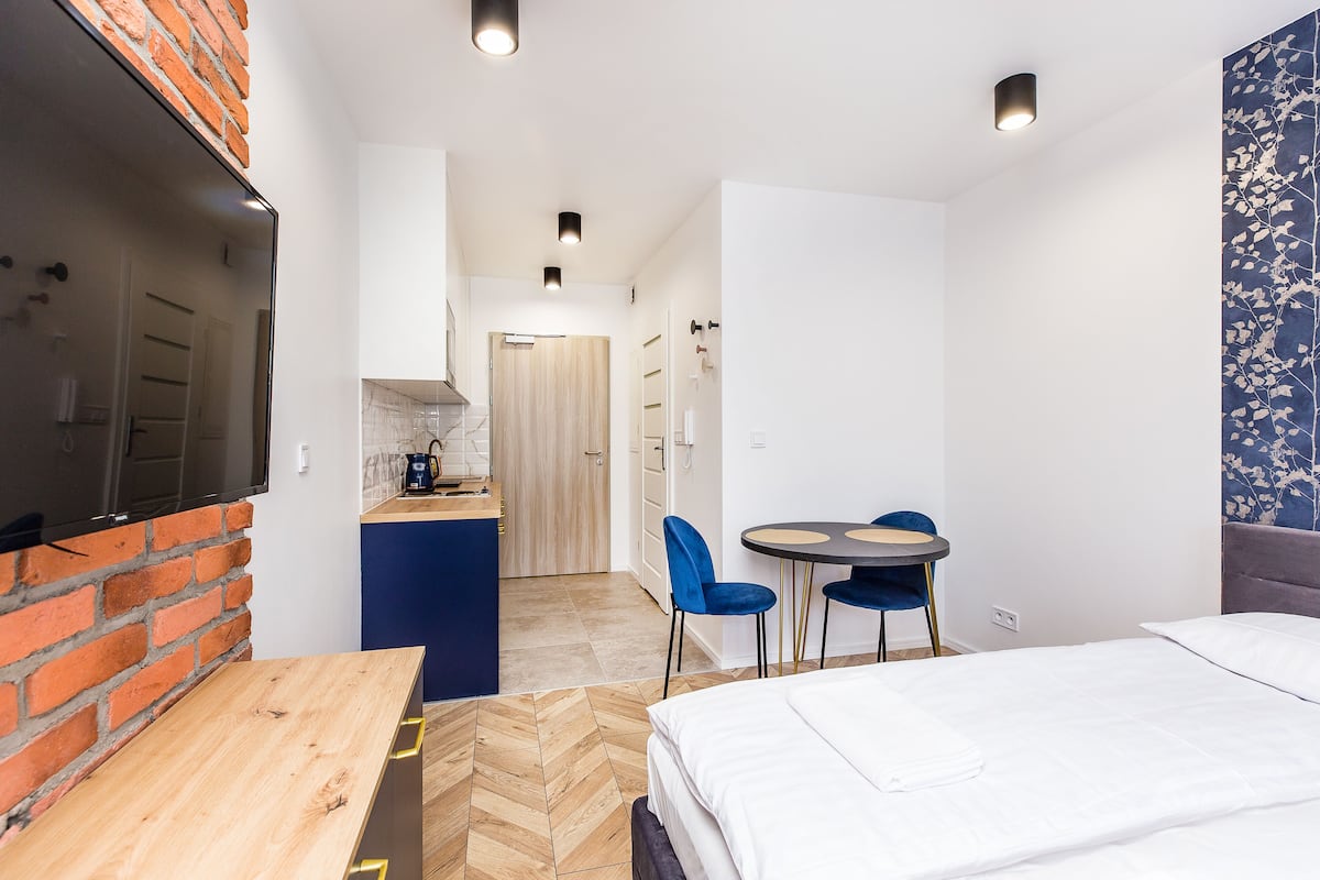 A compact hotel room with a bed, a wooden table, a small kitchen area, and a dining table with chairs.
