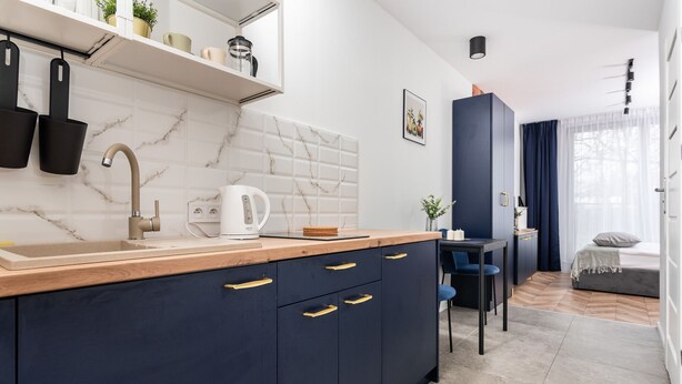 A modern kitchen with a marble backsplash, a coffee maker, and a sink. There is a dining area with a table and chairs, and a bedroom with a bed and a window. - APARTAMENTY NA DĘBNIKACH (Kraków)