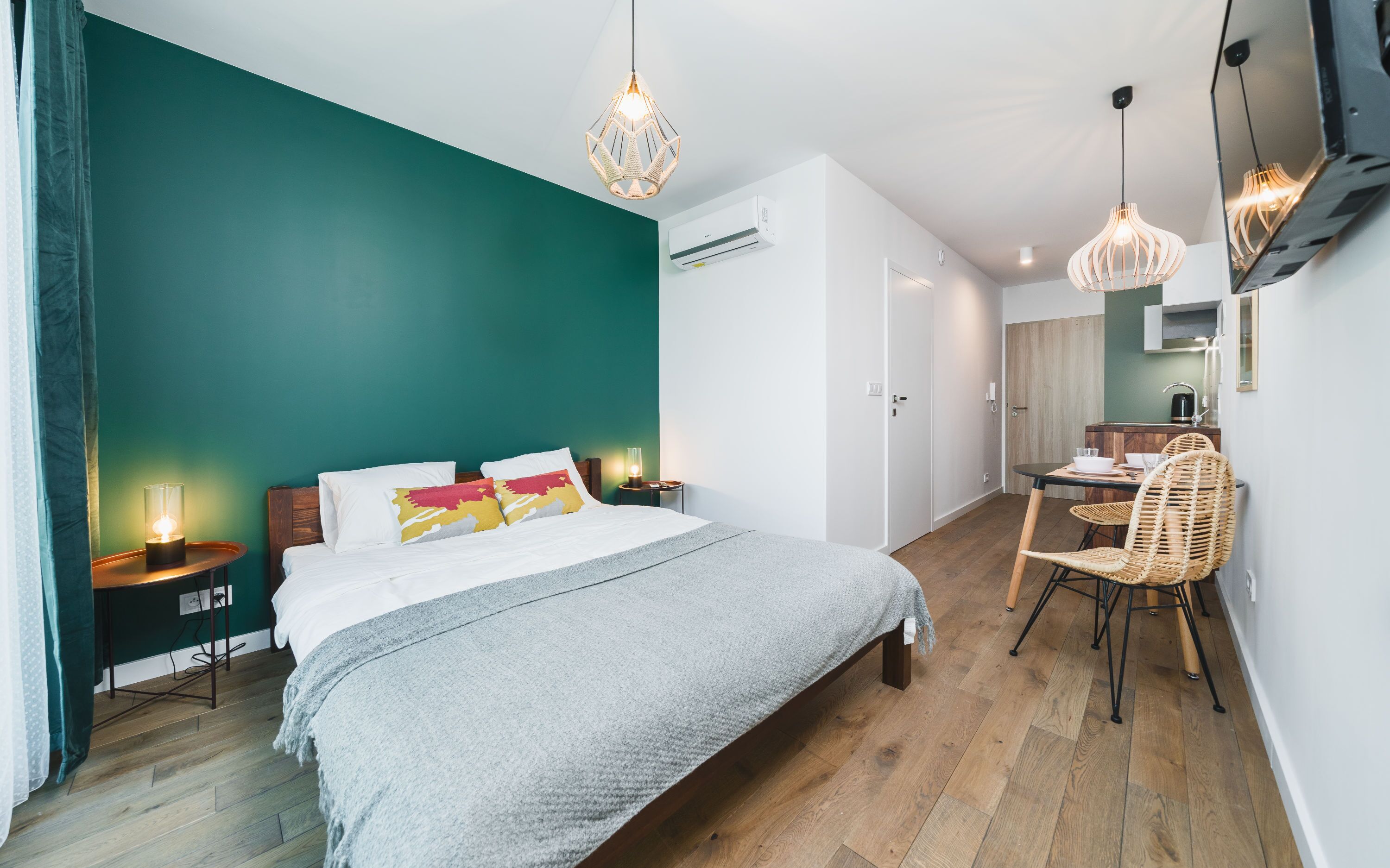 A modern bedroom with a teal accent wall, a bed with white and colorful pillows, a bedside table with a lamp, and a dining area with a wicker chair and table.