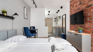A modern hotel room with a large bed, a desk, a chair, a TV mounted on a brick wall, and a small kitchenette in the background. - APARTAMENTY NA DĘBNIKACH (Kraków)