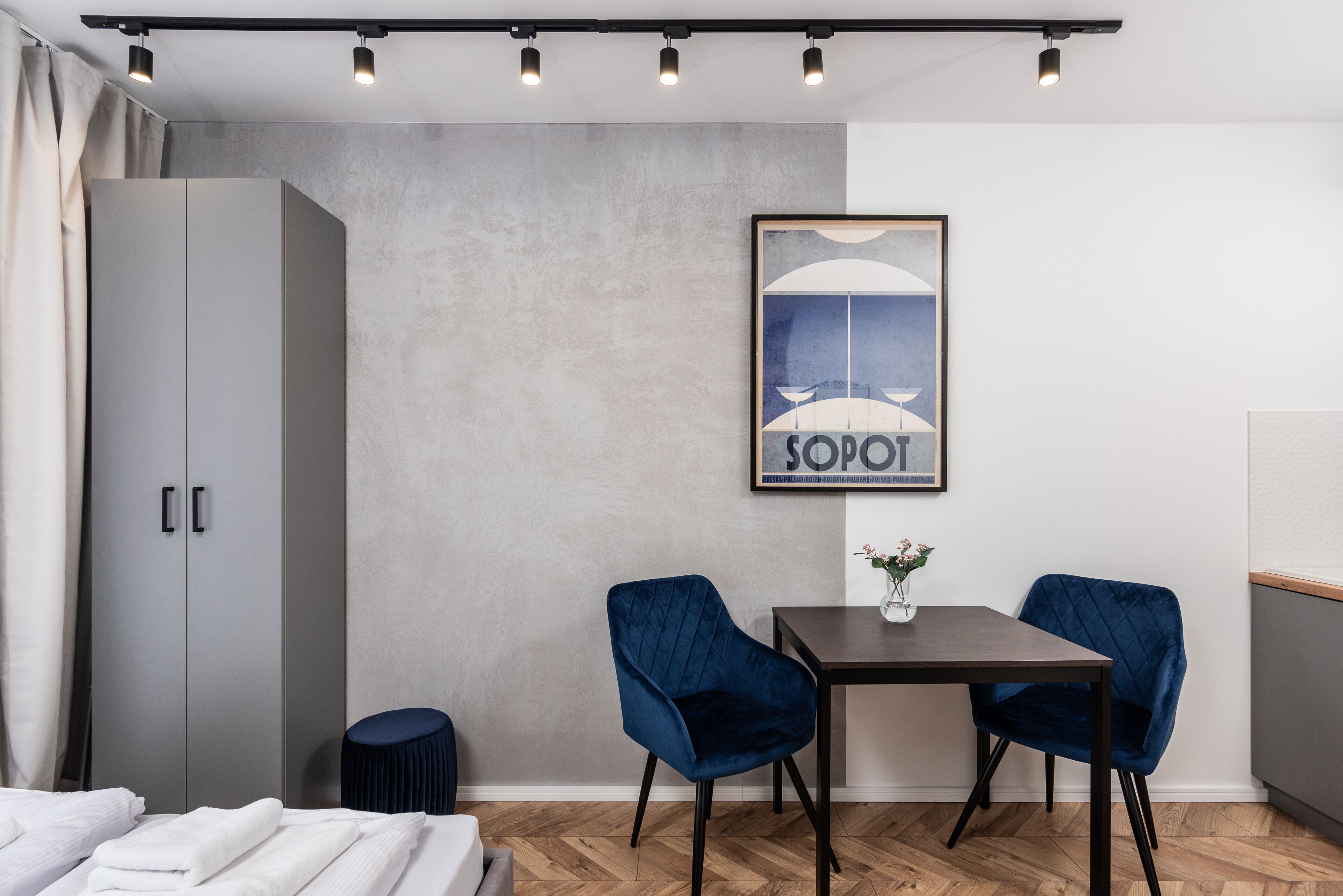 A modern hotel room with a grey wardrobe, a table with two blue chairs, a framed picture on the wall, and a wooden floor.