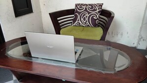 Ein Laptop steht auf einem Holztisch mit Glasplatte, ein brauner Stuhl mit gemustertem Sitzkissen und ein Fenster mit schwarzem Rahmen.