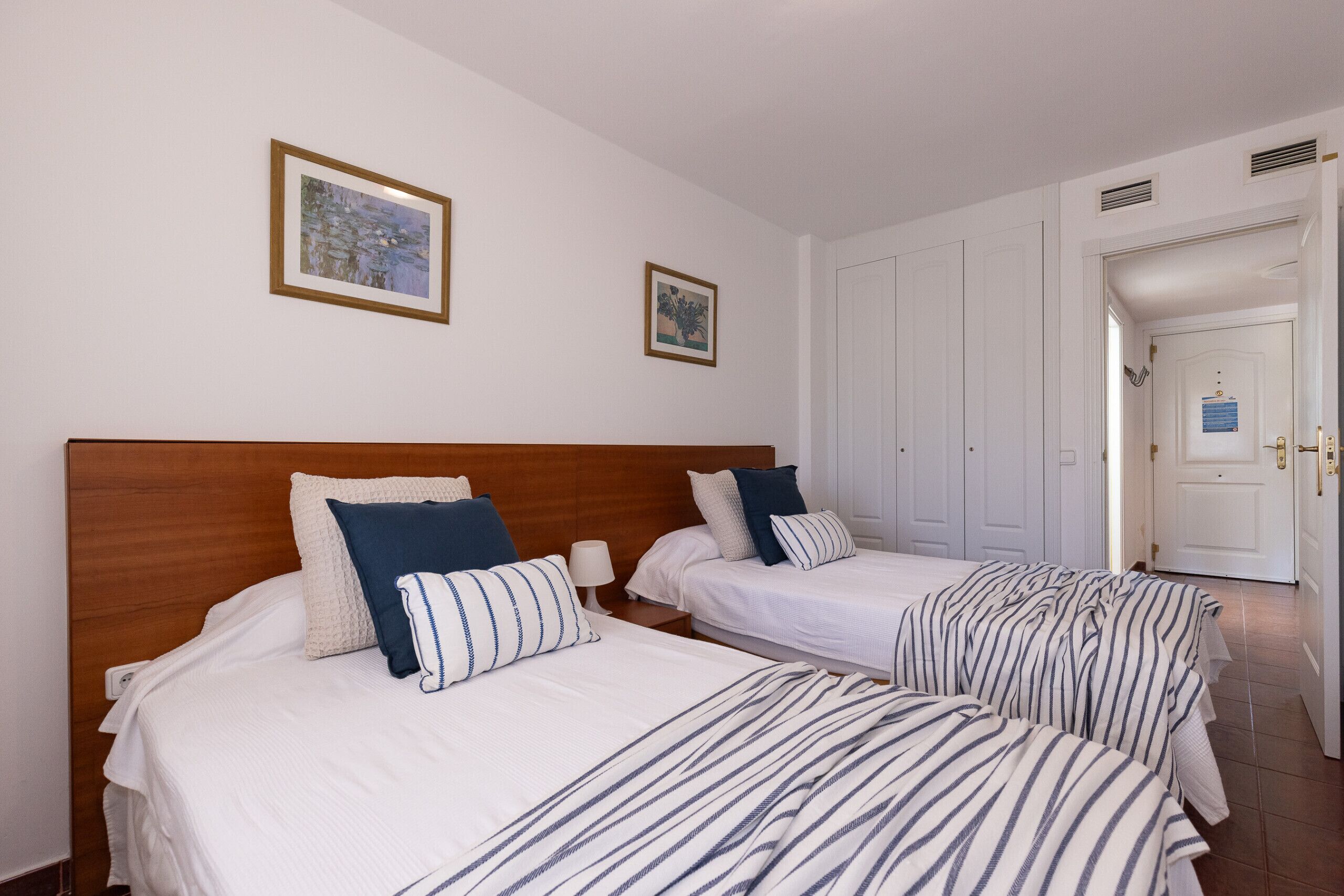 Een hotelkamer met twee bedden, elk voorzien van streepjesbeddengoed en kussens. Er hangen prenten aan de muur en een deur die naar een andere kamer leidt.