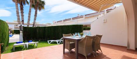 Un patio exterior con una mesa y sillas, una piscina al fondo y palmeras.