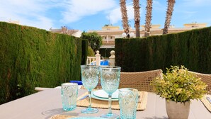 Une table dressée avec de la verrerie bleue, une plante en pot et une chaise en osier, dans un jardin offrant une vue sur un bâtiment et des palmiers.