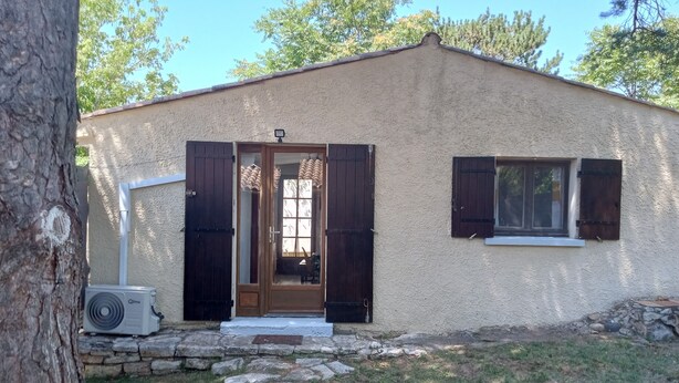 A single-story building with a beige exterior, a wooden door with dark shutters, a small window with shutters, and a stone pathway leading to the entrance. - Maisonnette Rustique (Le Garn)