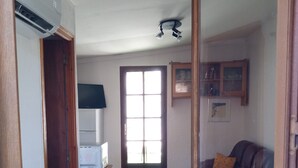 A living room with a sofa, a coffee table, and a dining table. There is a door leading to another room, and a window with blinds. - Maisonnette Rustique (Le Garn)