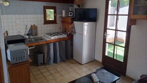 A compact kitchen with a gas stove, sink, and a refrigerator. There is a television mounted above the stove and a window with a view of the outdoors. - Maisonnette Rustique (Le Garn)