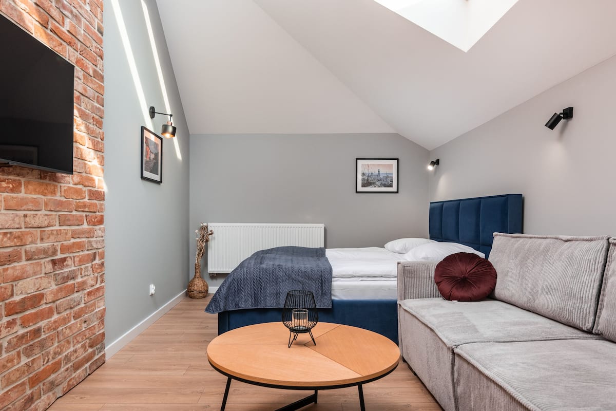 Quarto moderno com parede de tijolos, cama com cabeceira azul, sofá cinza e mesa de madeira redonda.