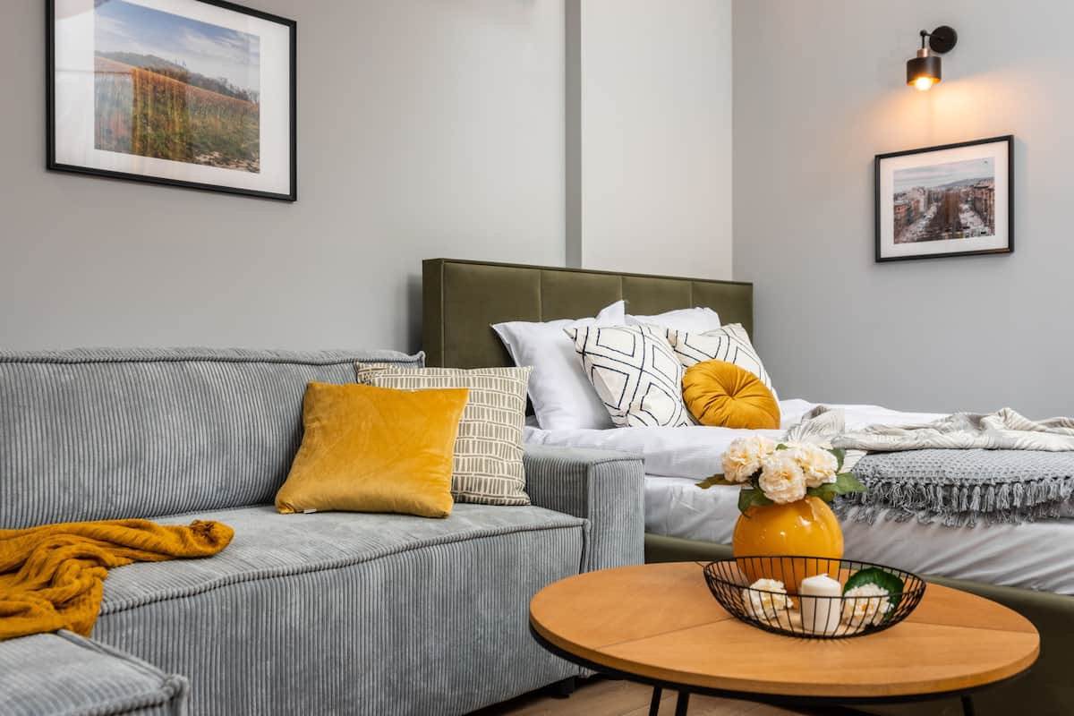 Um quarto moderno com um sofá cinzento, uma cama com roupa de cama estampada, uma mesa de cabeceira com um vaso de flores e duas imagens emolduradas nas paredes.