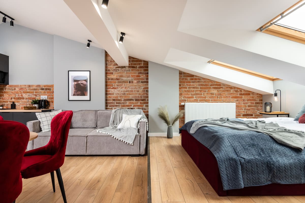 Quarto de loft moderno com cozinha no mezanino, uma cadeira vermelha, um sofá cinza e uma cama com colcha azul.
